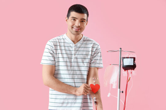 Young man with paper heart donating blood on pink background