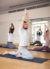 Fototapeta na wymiar Sporty young man practicing Hatha yoga with group in modern fitness studio, making stretching exercises..
