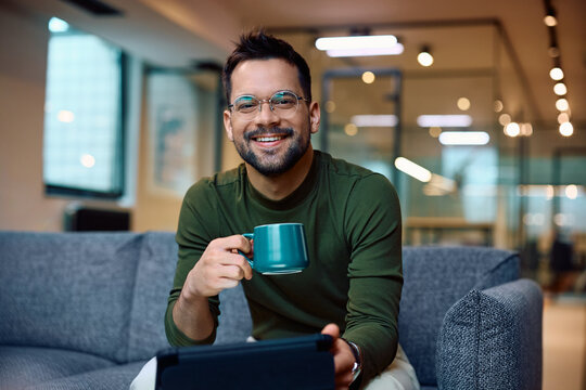 Happy entrepreneur drinks coffee while using digital tablet in office and looking at camera.
