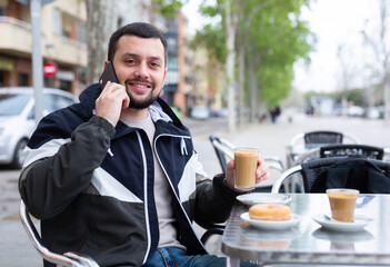 Cheerful young man traveler talking on phone while enjoying breakfast with warm cocoa and sweet soft donut at table