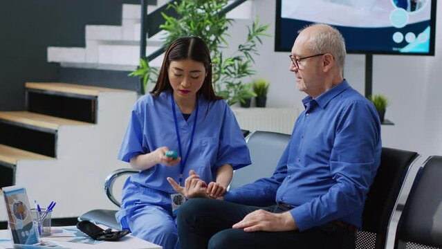 Asian nurse measuring sugar level with blood sample to do insulin test with glucometer for elderly patient with diabetes during examination. Assistant checking glucose in hospital waiting room
