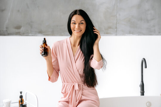 Split ends repair treatment. Female beauty routine. Haircare concept. Lovely brunette caucasian woman applying essential oil spray on her long hair while sitting in a bathroom, looks at camera, smiles