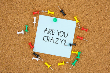 Are you crazy text on light blue post-it paper pinned on bulletin cork board surrounding by multi color pins. This message can be used in business concept about asking are you crazy.