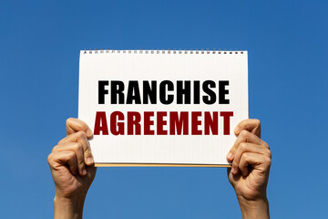 Franchise agreement text on notebook paper held by 2 hands with isolated blue sky background. This message can be used as business concept about franchise agreement.