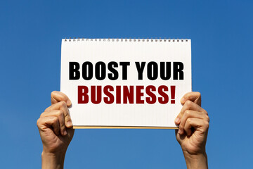 Boost your business text on notebook paper held by 2 hands with isolated blue sky background. This message can be used as business concept about boost your business.