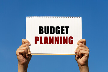 Budget planning text on notebook paper held by 2 hands with isolated blue sky background. This message can be used as business concept about budget planning.