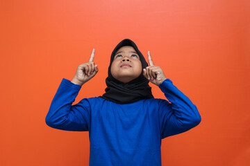 Excited Asian muslim girl wear hijab pointing fingers up showing empty space isolated orange color...