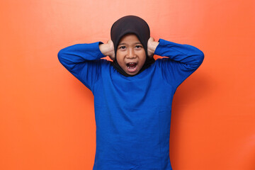 Asian Muslim little girl screaming covering ears with both hands, distressed by noise isolated over orange background