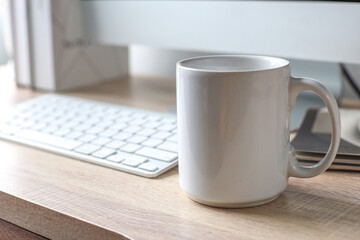 Blank mockup white coffee mug on office table near computer and window with daylight. 
