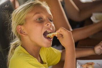 Adorable curly blonde boy eating pizza at the camera in italian restaurant. Portrait of child with long blonde hair and a yellow T-shirt eats junk food in a cafe. Blur on the background.
