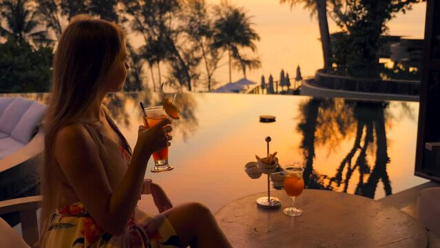 Young woman holds cocktail in outdoors bar at luxury hotel in summer. Attractive girl in elegant dress with mocktail at evening in cafe at tropical resort. Travel, tourism, holiday, vacation concept.