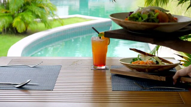 In a tropical setting, a waiter carefully arranges the table at an outdoor restaurant. With the shimmering pool and swaying palm trees in the background