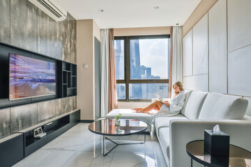 Young woman on vacation sitting on sofa with laptop in a luxury hotel modern room. Relaxing in a white bathrobe, she sits on a couch near the window. Self-employment and remote work.