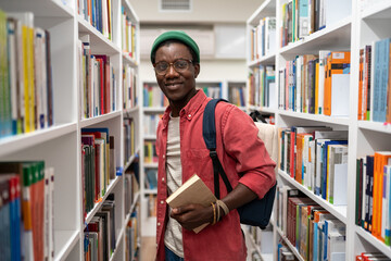 Smiling african american man student holding book in university library standing between...