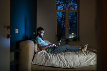Black student guy lying in bed with laptop in evening, studying until late hours. African American remote employee working overtime from home, doing freelance work at night
