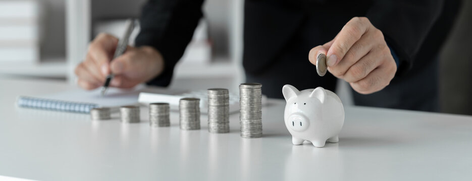 Image of businessman and pile of money showing growth Put the coins in the piggy bank and record the income. expenses on desk money saving concept business finance investment planning.