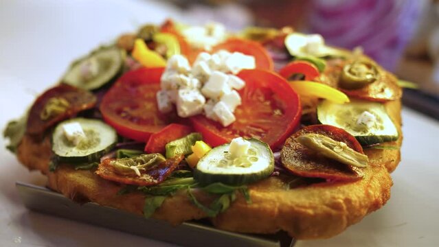 Langos Topped With Sliced Tomatoes, Cucumber, Bell Pepper, And Cheese Served On The Counter. - zoom out