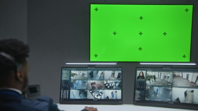 African American male operator watches CCTV cameras with AI face scanning system in security control center. Computer monitors showing surveillance cameras footage. Big digital screen with chromakey.
