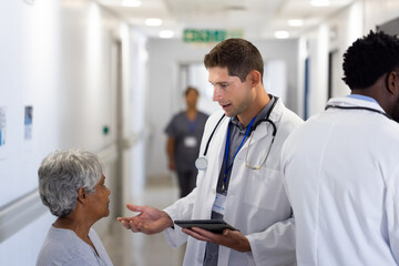 Caucasian male doctor with tablet talking with diverse senior female patient in busy corridor