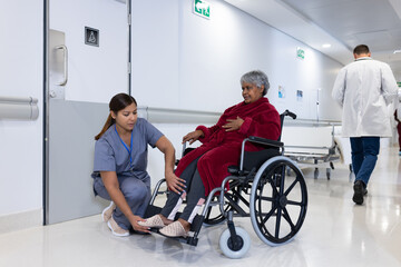 Asian female doctor checking leg of diverse senior female patient in wheelchair in hospital corridor