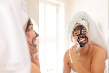 Plus size caucasian woman with towel on removing beauty face mask looking in bathroom mirror smiling