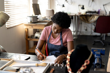 African american female designer at desk sketching designs in notebook at jewellery workshop