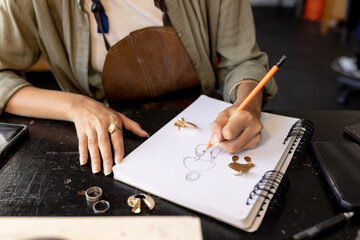 Midsection of biracial female jewellery designer at desk sketching designs at workshop