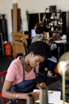African american female designer sketching designs in notebook at jewellery workshop, copy space
