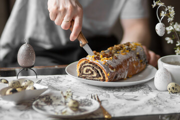 Man cutting poppy seed roll with knife. Festive table with poppy seed Easter cake, quail eggs, candles, spring branches.