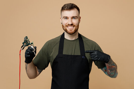 Smiling tattooer master artist tattooed man wearing green t-shirt apron hold point index finger on machine black ink in jar, equipment for making tattoo art on body isolated on plain beige background.