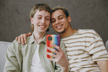 Young happy couple two gay men wear casual clothes together hold use mobile cell phone sit on sofa couch at home flat rest spend free spare time in living room Pride day june month love lgbtq concept - obrazy, fototapety, plakaty
