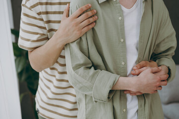 Close up cropped young couple two gay men wearing casual clothes together hug cuddle stand near window at home flat rest spend free spare time in living room. Pride day june month love lgbtq concept.