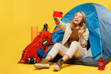 Full body young woman sit near bag with stuff tent do selfie shot mobile cell phone show v-sign isolated on plain yellow background. Tourist walk on spare time. Hiking trek rest travel trip concept.