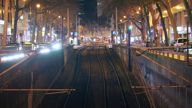 Timelapse with symmetrical arrangement of trams and traffic at the train station. Trams smoothly enter and exit while cars flow on the side. Tracks in the foreground provide a visual focal point. zoom