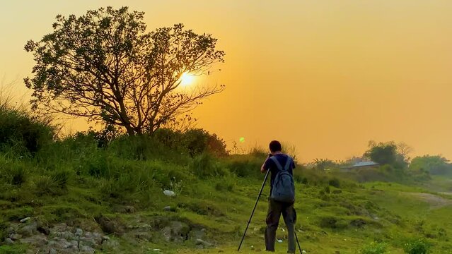 Photographer with tripod composes shot above yellow sunset sky and tree on hill