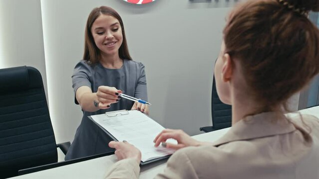 Selective focus shot of young receptionist working at check-in desk in modern clinic explaining patient how to sign document