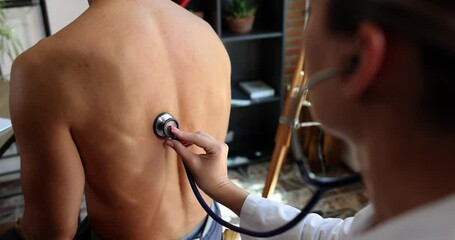 Female doctor checks lungs of man with stethoscope in clinic. Woman puts medical equipment to bare back of male patient at appointment slow motion