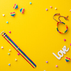 Show your solidarity with this top square view flat lay of LGBT parade accessories on a yellow background, including rainbow wristlets, pin badges, and hearts, with an empty frame for text or advert