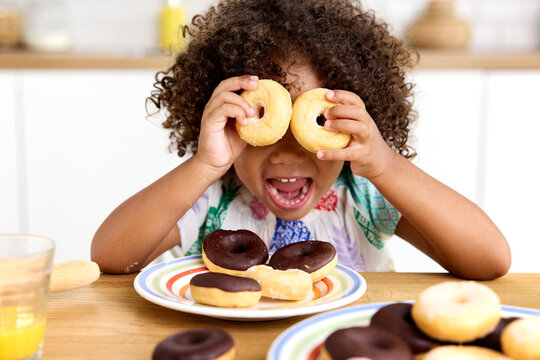 Funny toddler girl at kitchen table holding donuts on her eyes