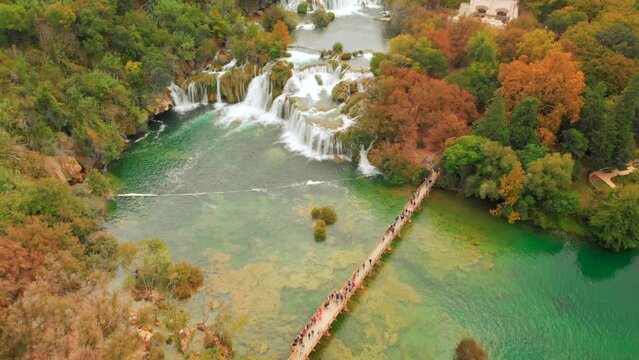 A drone captures the Krka waterfalls in stunning detail. People cross a wooden bridge in the foreground