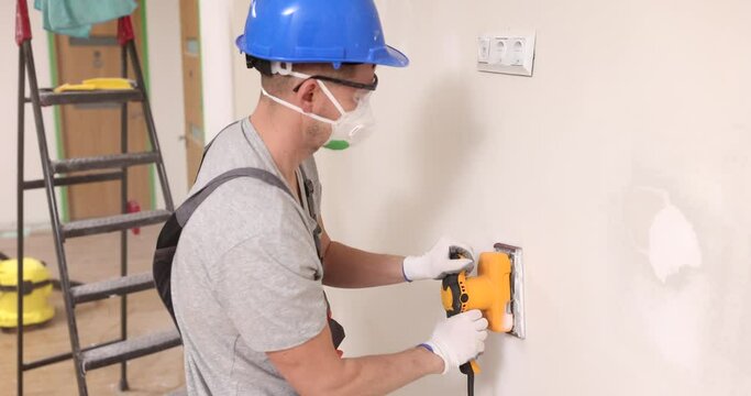 Worker with respirator polishes drywall with electric sander in room slow motion. Builder grinds surface after plaster applying in enclosure