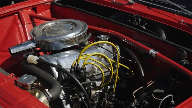 Clean and shiny old carbureted engine in engine compartment of red old car. Under sunlight is demo retro car without hood. Passing people are reflected in chrome air intake.