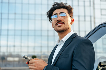 Young businessman standing near luxury car and using smartphone