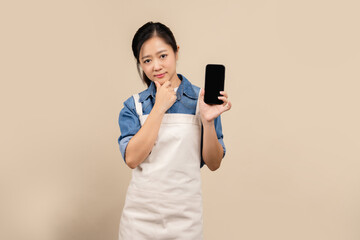 Naklejka premium Young Asian waitress wearing apron. She had a question with a curious expression on her face and was holding her smartphone with a blank screen. Business problem and solution concept
