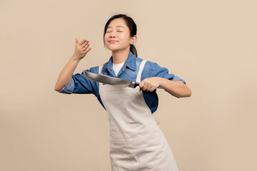 Young Asian housewife in apron and holding pan standing isolated on light brown background. She was...