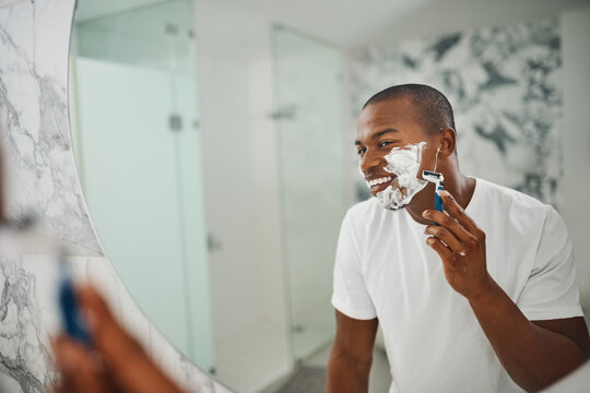 Black man, smile and shaving with razor, foam or grooming in mirror for self care in home bathroom. African guy, soap or cream for facial hair removal, beard or clean for hygiene in morning at house - Powered by Adobe