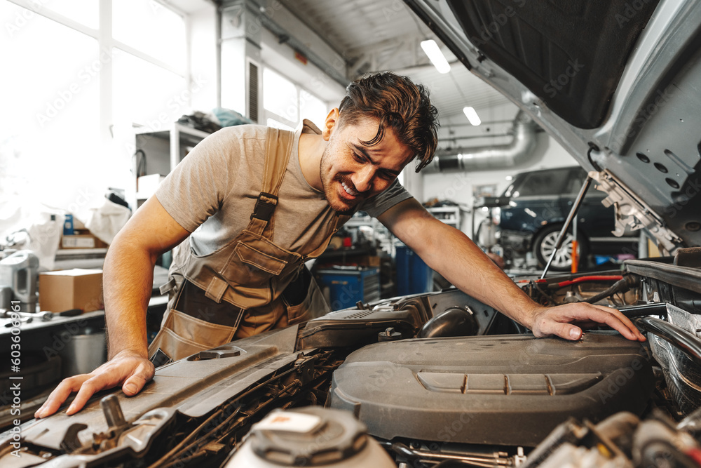 Wall mural Young male mechanic examining engine under hood of car at the repair garage