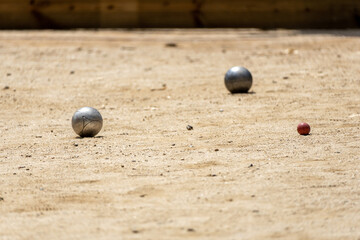 Two metal balls from the game of petanque approaching the bowling ball on a sandy ground of a petanque court on a sunny day with the shadows of the trees on the ground