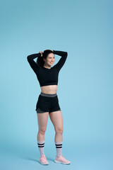 Young smiling strong fitness woman wearing black bra and sports shorts, standing over blue backdrop