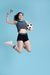 Athlete woman smiles, jumps high up over blue background, holds soccer ball and golden champion cup